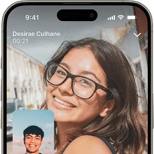 A video call interface on a phone. The main screen shows a smiling woman outdoors with large glasses. In the bottom left corner, there is a small window showing a man’s face, indicating the caller.
