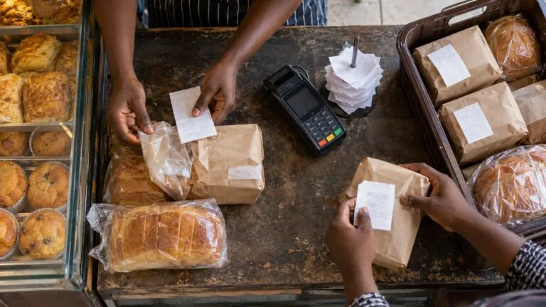 Overhead view of bakery staff matching packaged orders with payment slips beside an unbranded card terminal on a worn counter.