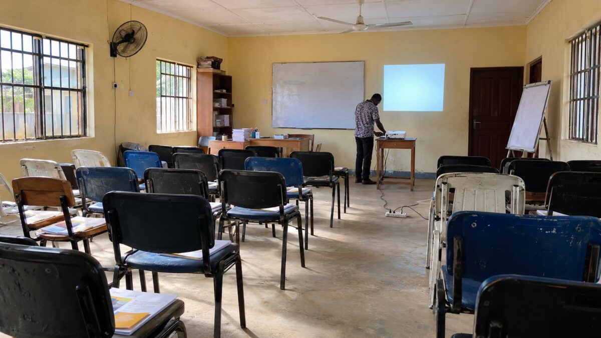 Wide view of a modest Nigerian training room with rows of chairs and an instructor testing a projector before a course session.
