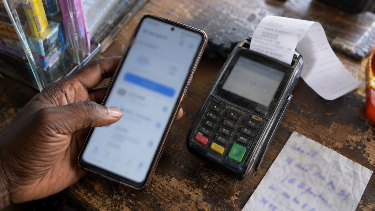 A hand holds a smartphone beside a worn card terminal, curled receipt and handwritten order slip on a shop counter.