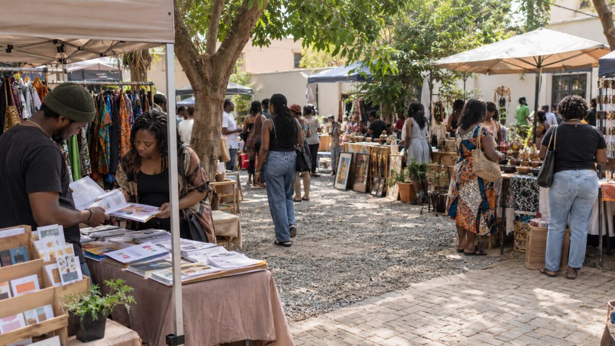 Wide view of a shaded Abuja creative market where two people review printed digital product samples at a small stall amid shoppers and craft vendors.